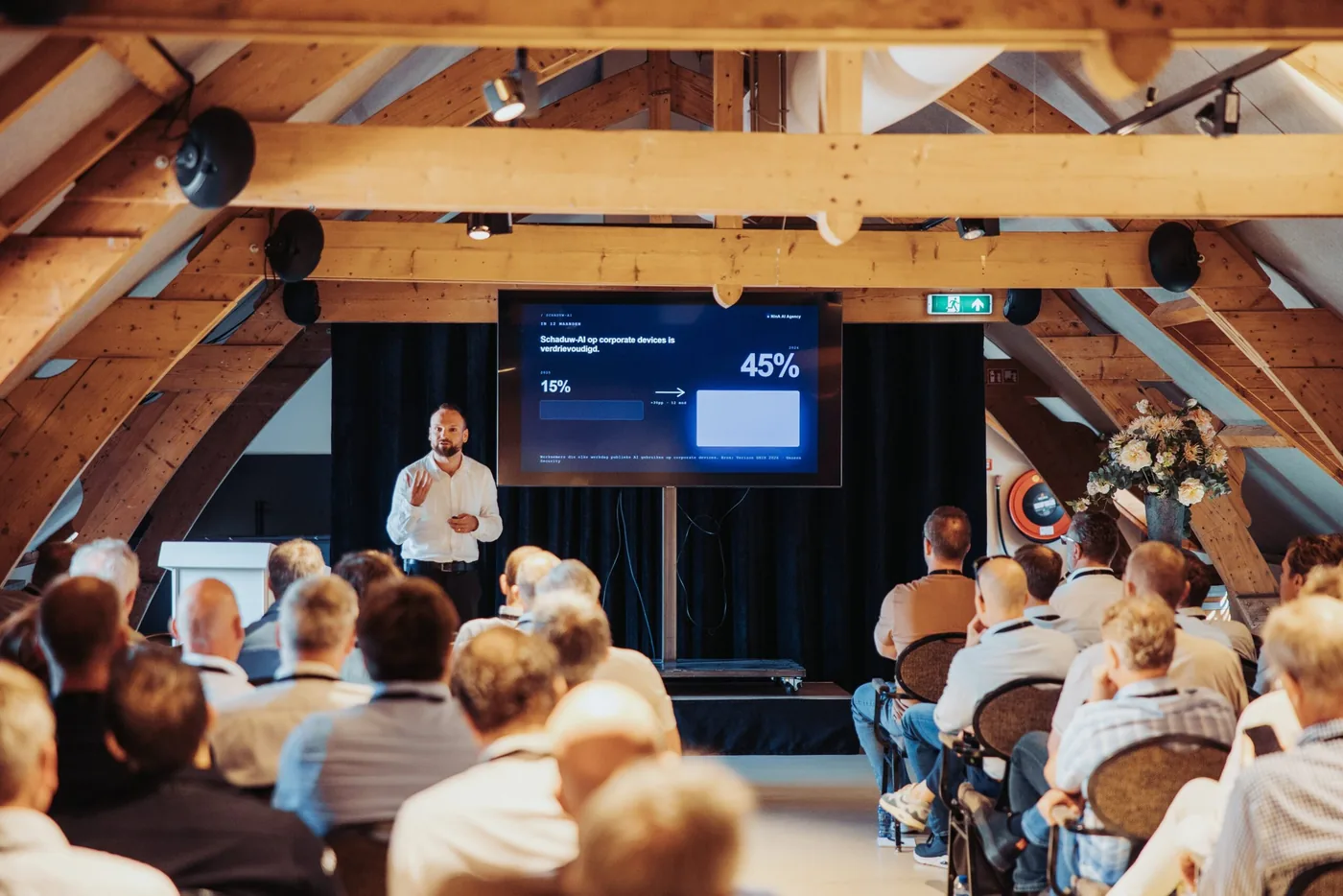 AI-keynote voor een zittende zaal onder een houten kapconstructie, met een slide over schaduw-AI op corporate devices