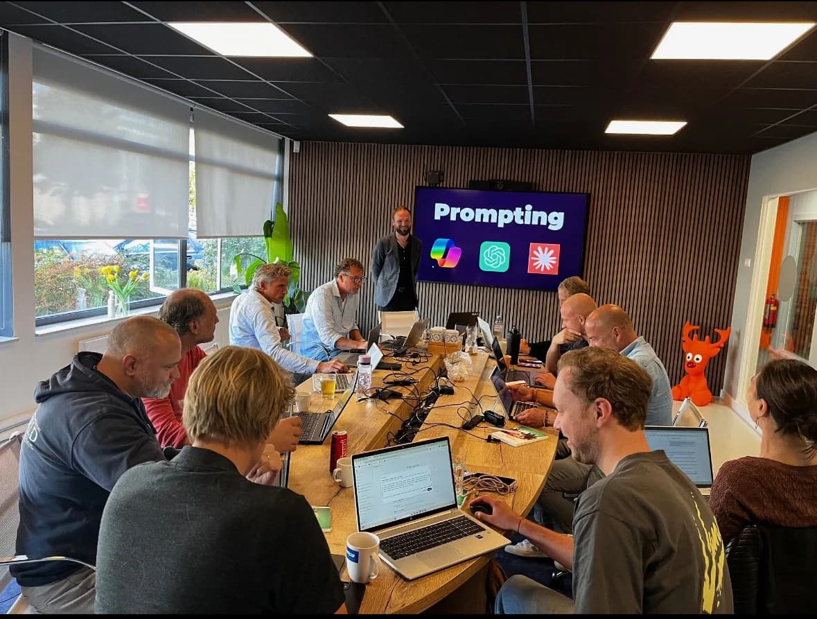 Deelnemers aan een hands-on AI-workshop achter hun laptop rond een grote tafel, met Prompting op het scherm